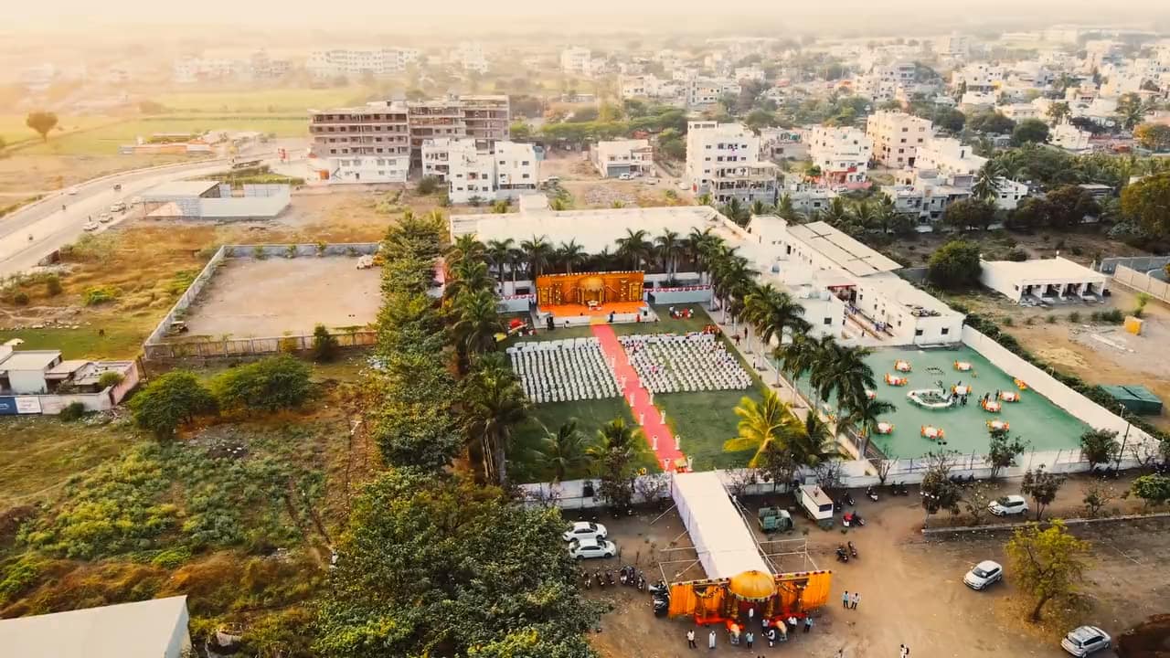 From a drone at golden hour: an open-air wedding venue, the mandap at the head of a red-carpet aisle between rows of chairs