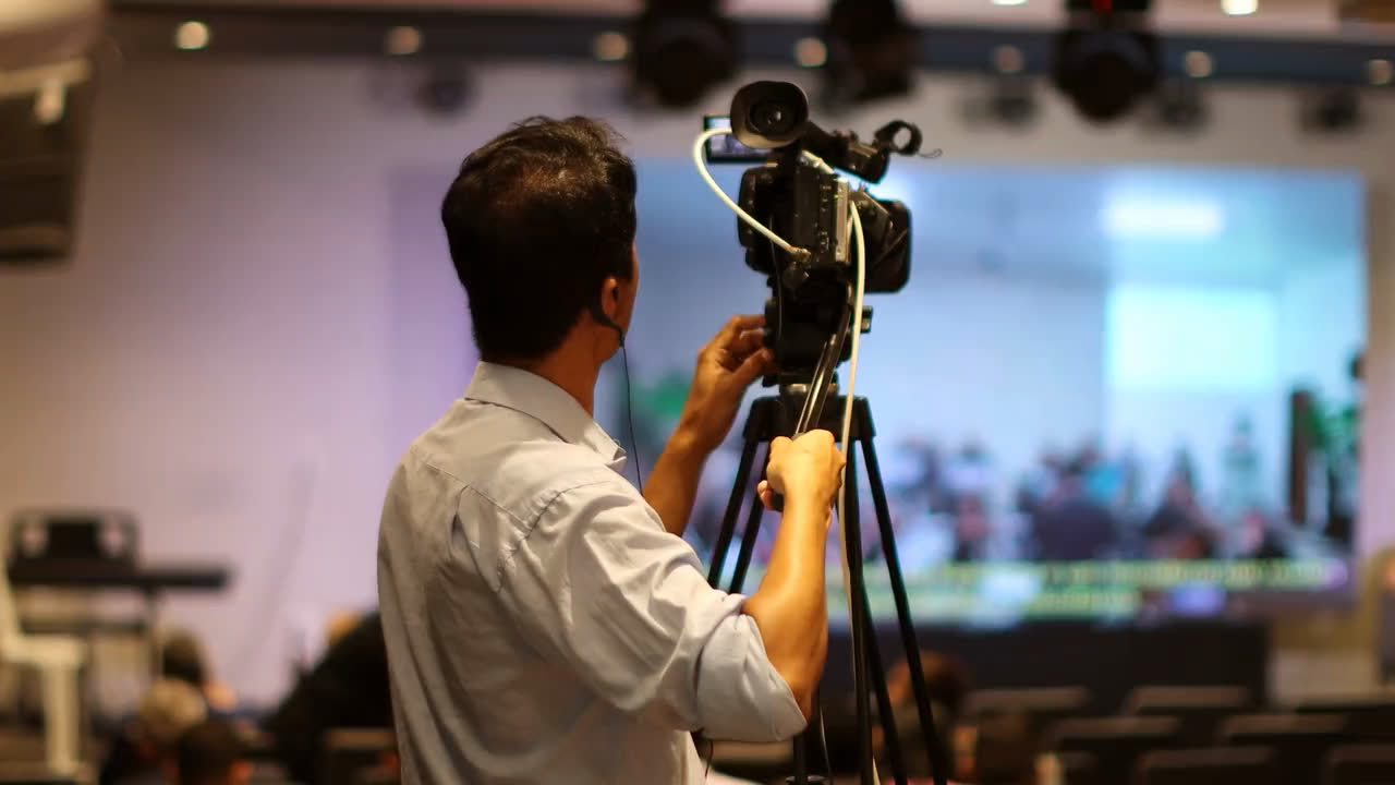 A camera operator on a tripod filming an event in a hall