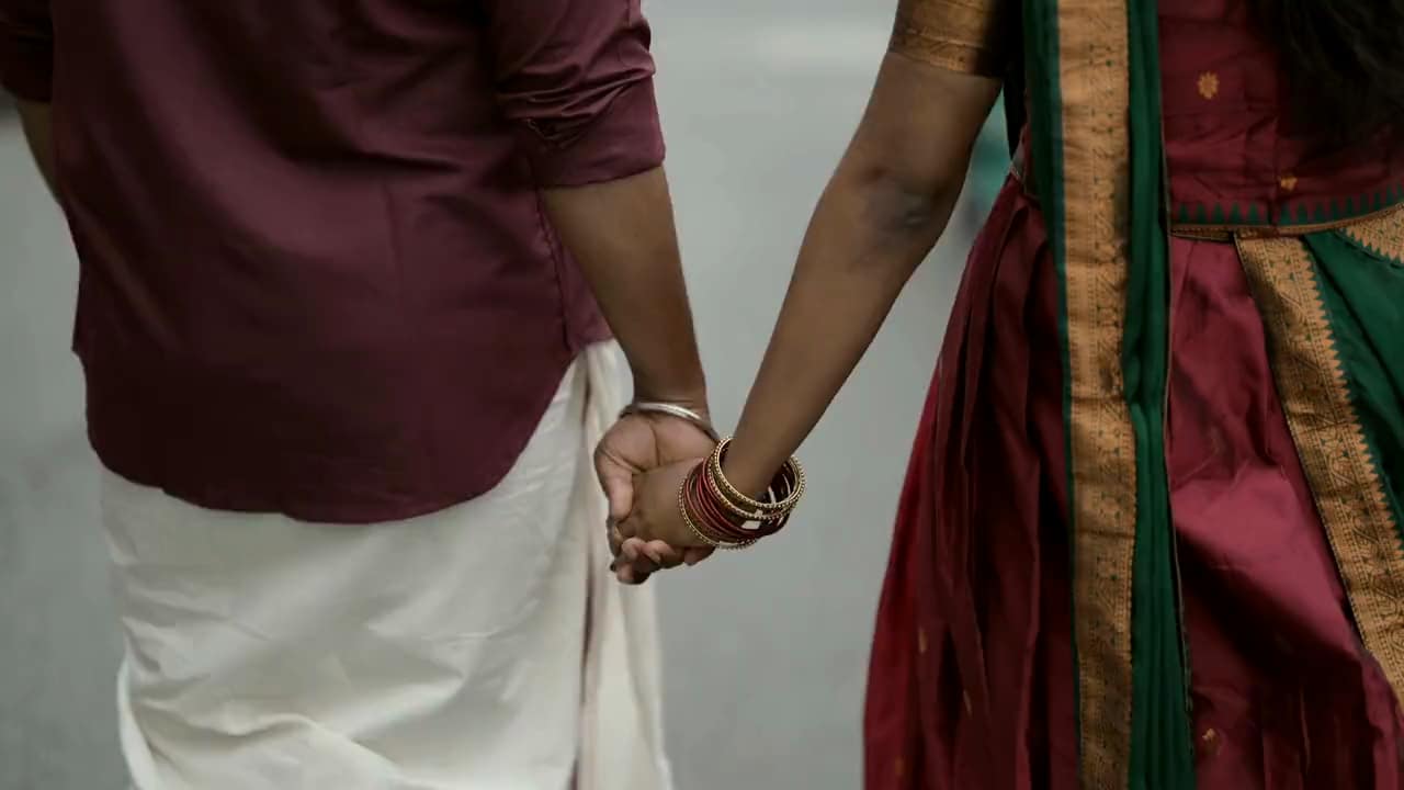 A couple walking hand in hand, close on their hands: he in a white dhoti and a maroon shirt, she in maroon silk with a gold and green border