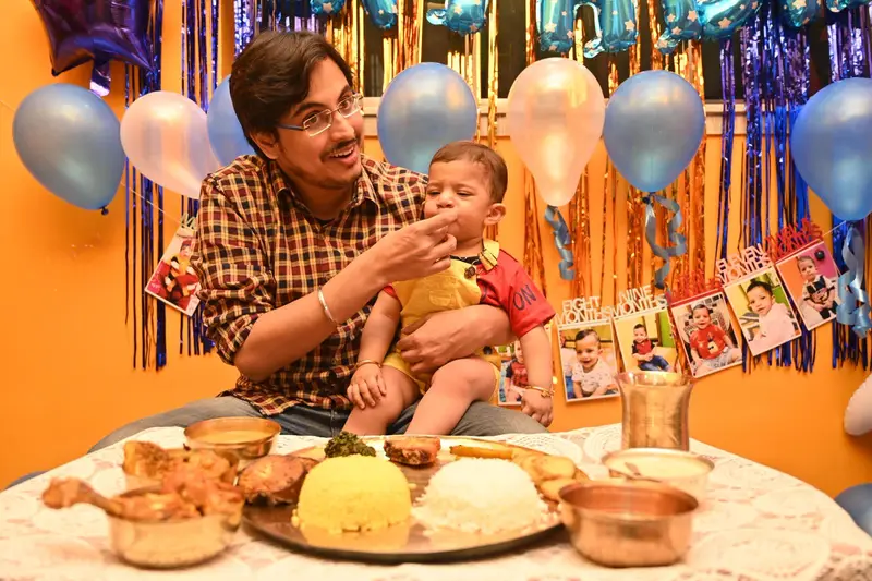 A father feeding his baby at a first-birthday table, balloons behind.