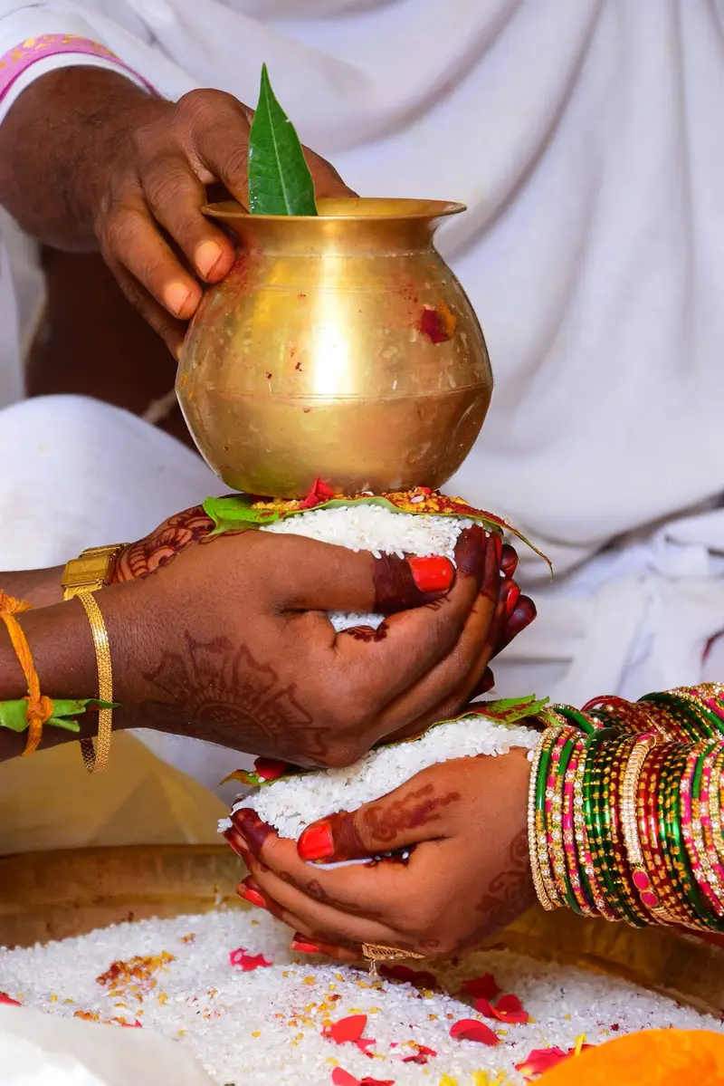 hands holding a brass kalasham of rice with mango leaves